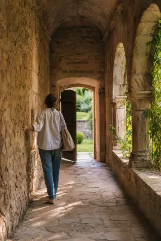 A person quietly exploring a sunlit stone corridor at chartreuse Val-de-Bénédiction, touching the wall, with greenery outside and a peaceful, contemplative mood.