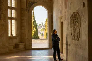 Woman quietly observing medieval frescoes in a softly lit historic hall, with Villeneuve-lès-Avignon rooftops visible through an arched window in the background, golden hour ambiance.