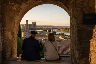 Two people quietly talking under a stone archway in Villeneuve-lès-Avignon at sunset, with soft sunlight illuminating historic rooftops and the landscape beyond.