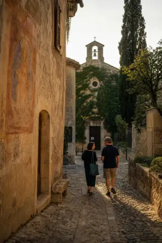 Two locals walk past a small ivy-covered chapel in a sunlit medieval Provençal village street, bathed in warm evening light.