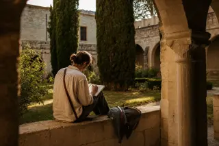 Traveler quietly sketching in a medieval cloister garden at sunset, warm light casting shadows on ancient stone architecture.