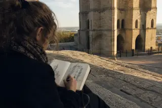 A person quietly sketching in a notebook on stone steps beside the Philippe-le-Bel tower, bathed in soft late-afternoon sunlight.