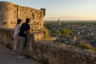 Person seated on stone wall in cloister courtyard at sunset, observing distant community activity among medieval arches