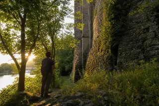 Hiker standing quietly in dappled forest sunlight, contemplating a mossy stone wall near a riverbank in golden hour light.