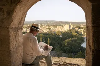 Older man quietly sketching Villeneuve-lès-Avignon from the shadowed archway of Fort Saint-André, sunlit rooftops and olive trees in the background.