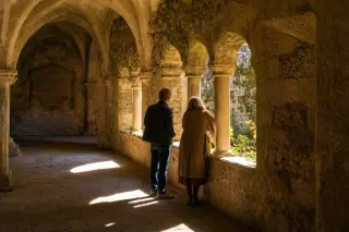 Elderly locals on a stone bench studying a map near a historic chapel in soft golden hour light, facing away from the camera, with subtle focus on their hands and the ancient stones nearby.
